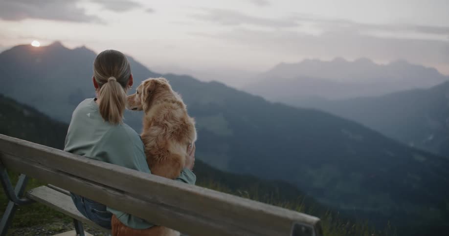 A woman and a dog sitting side by side on a bench overlooking a mountain range during twilight
