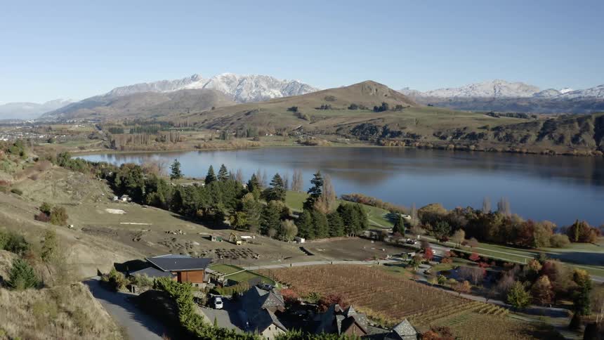 Cars drive past a rural area near a calm lake and snow capped mountains