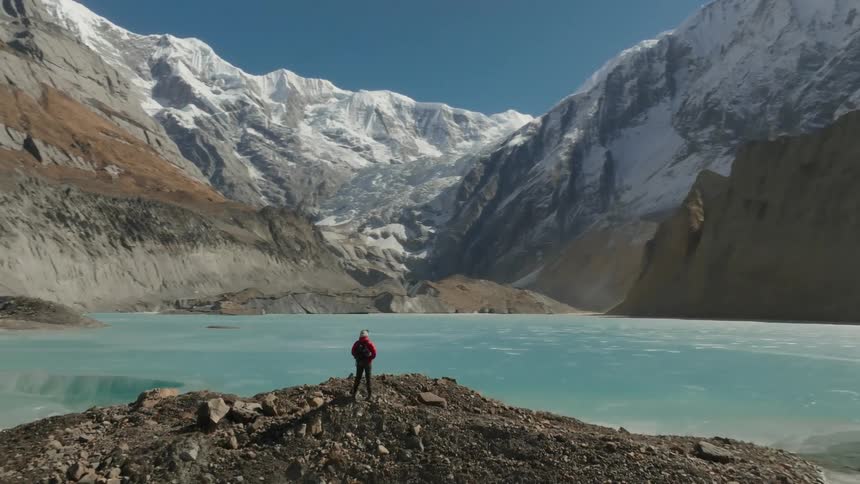 A person standing on a rocky outcrop gazing at a glacial lake surrounded by snow capped mountains