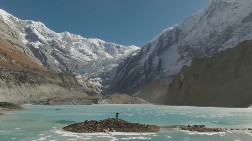 Person with open arms in the center of a rock surrounded by a blue lake and snowy mountains