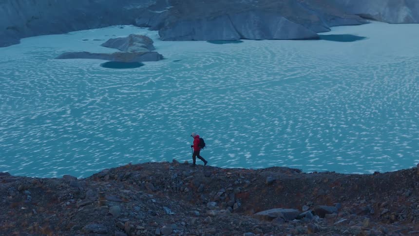 Aerial view of a person in a red jacket hiking on a mountainous cliff at the edge of a body of water