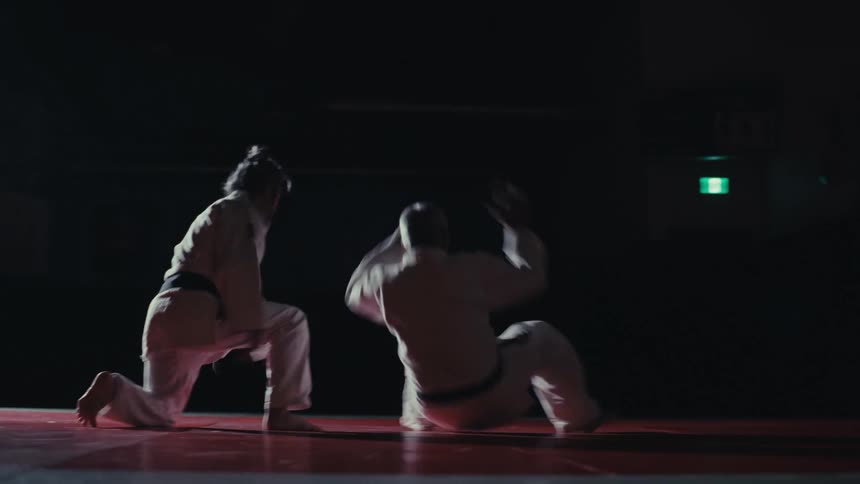 Two male martial artists compete on tiled floor in a large dark room