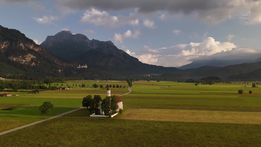 A small church with a steeple nestled amidst expansive green fields framed by mountains under a partly cloudy sky