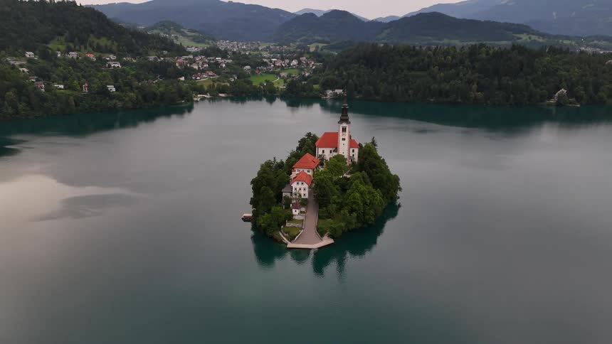 Aerial scene of a church on an island in a lake surrounded by a valley and cities in the background