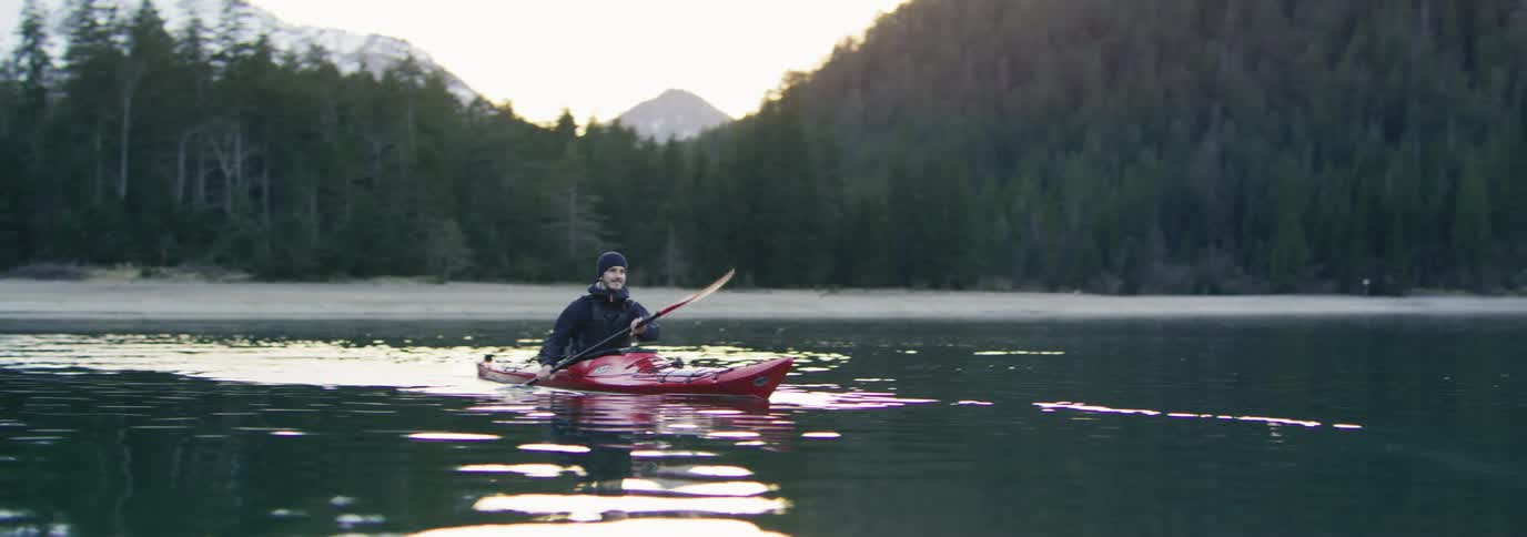 Man paddling a red kayak in calm waters of a lake with mountains around