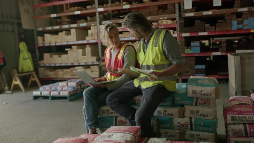 Two workers wearing identification vests sitting on loads talking to each other with documents and a laptop