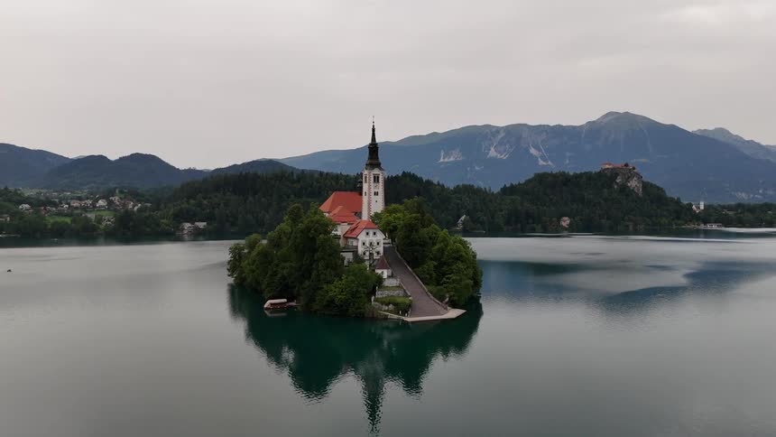 Lake water reflects a church standing on a small island across from a town on the forested shore