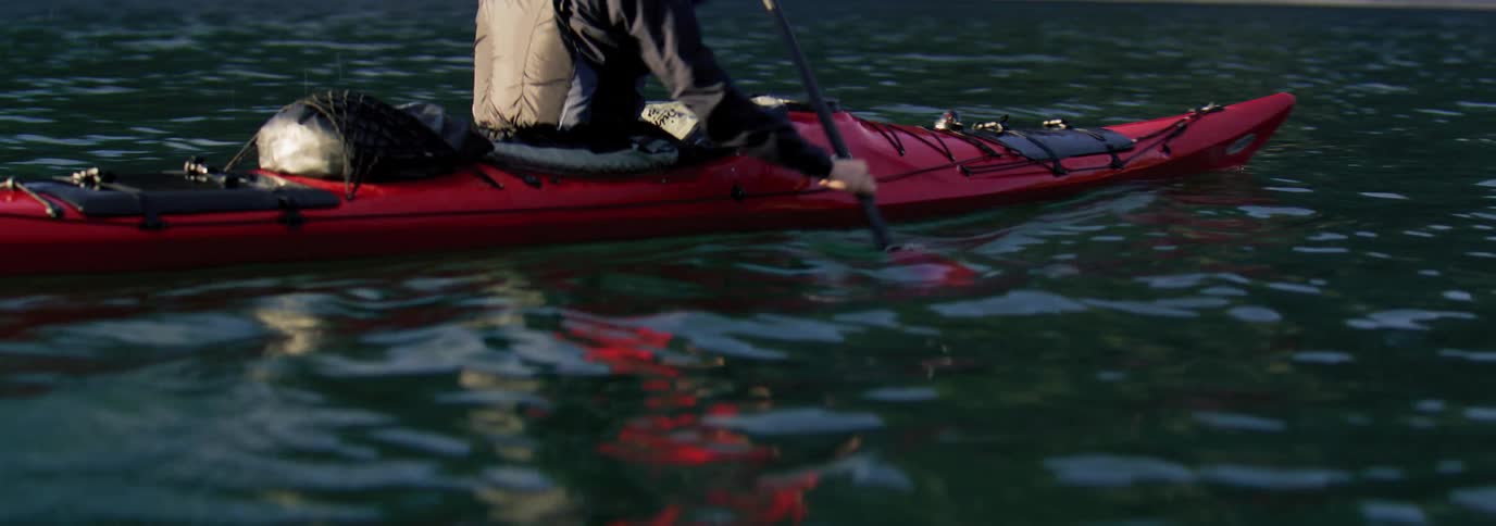 Man in a calm sea riding a red kayak with mountains around