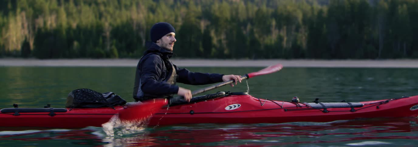 Man moves kayak through the lake in front of the forest and mountains using his oar
