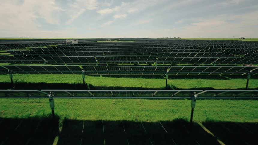 Multiple rows of solar panels sit in the grass in a field under a summer sky