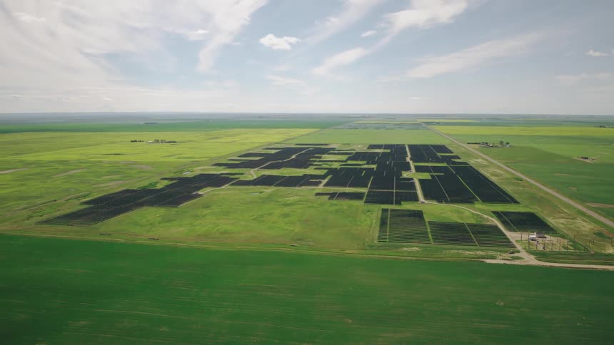 Solar panels partially fill a grassy field surrounded by farms
