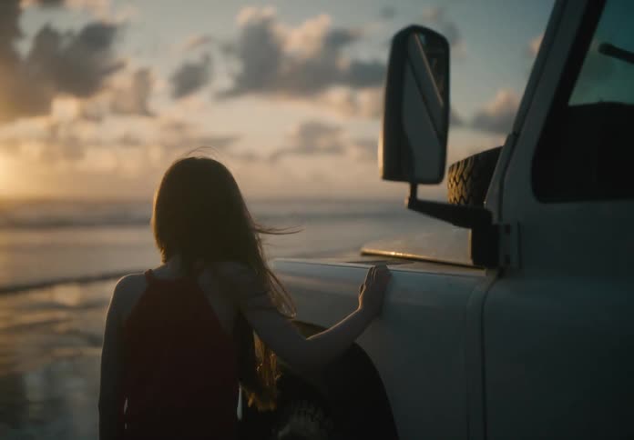 A teenage girl leaning against the door of a parked vehicle gazing toward a sunset over the ocean