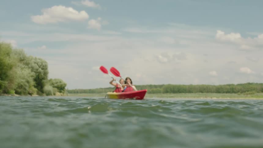 Two people paddle a red kayak on a serene lake surrounded by lush greenery and a clear blue sky.