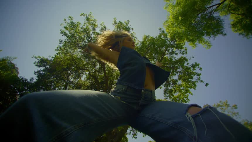 A woman dances outdoors wearing denim with sunlight filtering through tree leaves