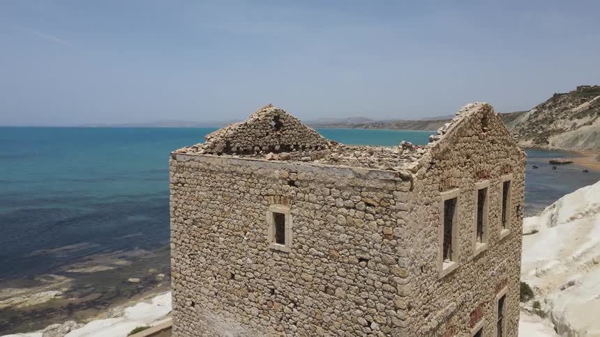 A historical roofless house built from rocks stands on a beach near the ocean