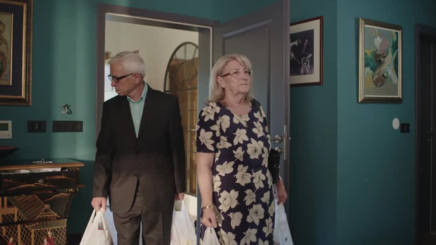 An elderly couple enter a room both looking forward with a thoughtful expression