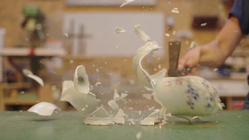 A hand uses a hammer to smash a porcelain teapot to pieces