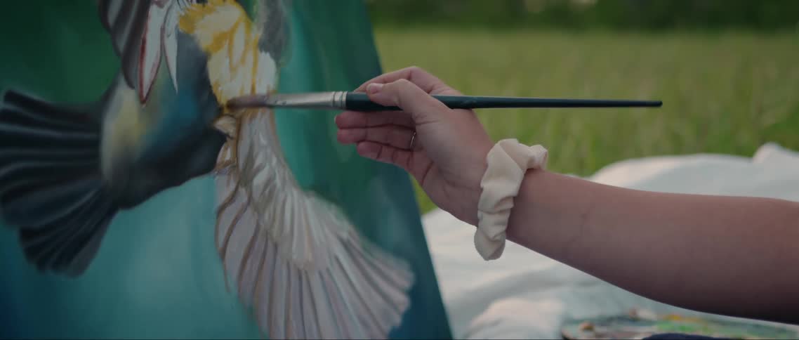 The arm of a person who is using a long brush to paint a bird in a colorful painting