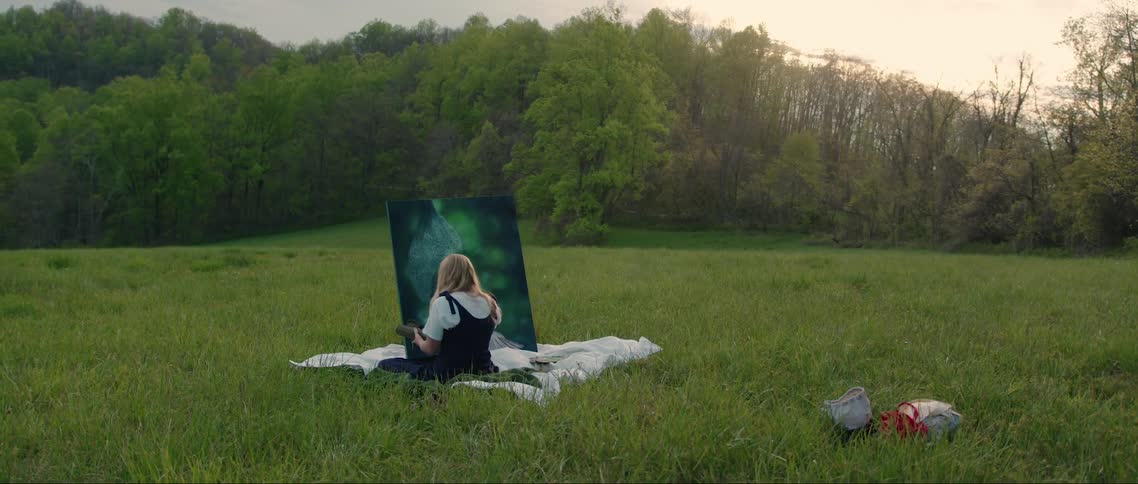 A woman sitting on a white blanket in a grassy field facing away from the camera towards a large canvas