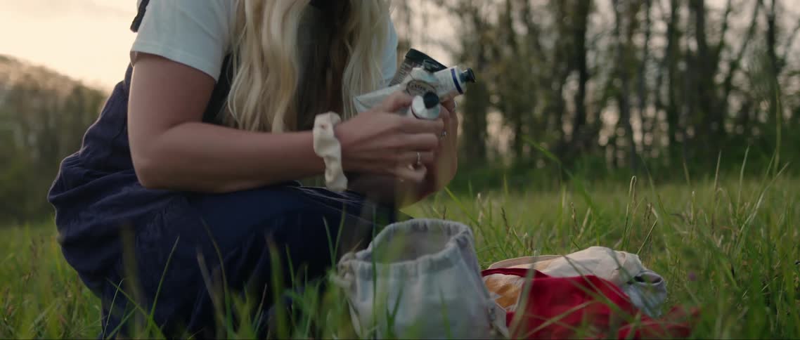 A blonde woman kneeling in a grassy area takes painting supplies from a small bag