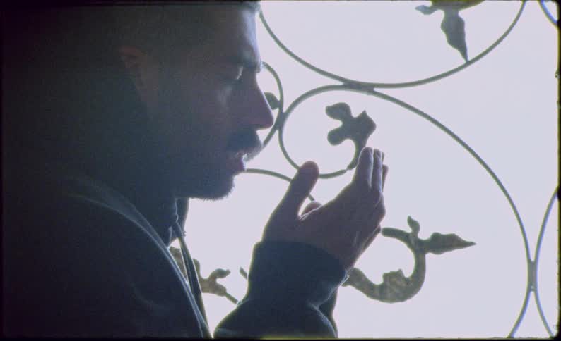 A monk in profile silhouetted against an ornate window engaged in prayer