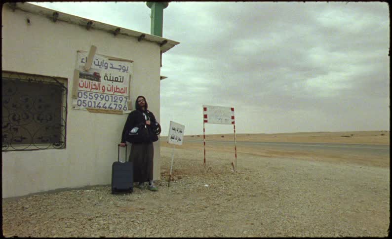 A person stands next to a small building in a desert area holding a suitcase and a sign with arabic text in the background