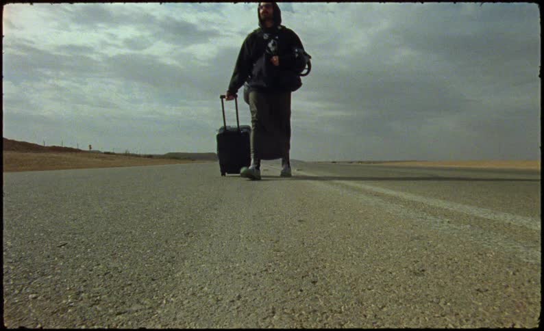 Man walking with a suitcase on a paved road in a desert environment