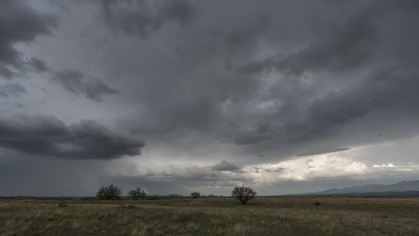 The dark clouds are rolling across the field that has a few sparse bushesthere is a mountain range to the right that slowly moves from sight