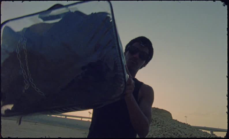 Man holding a transparent suitcase with clothes inside with a sunset in the background and an overpass