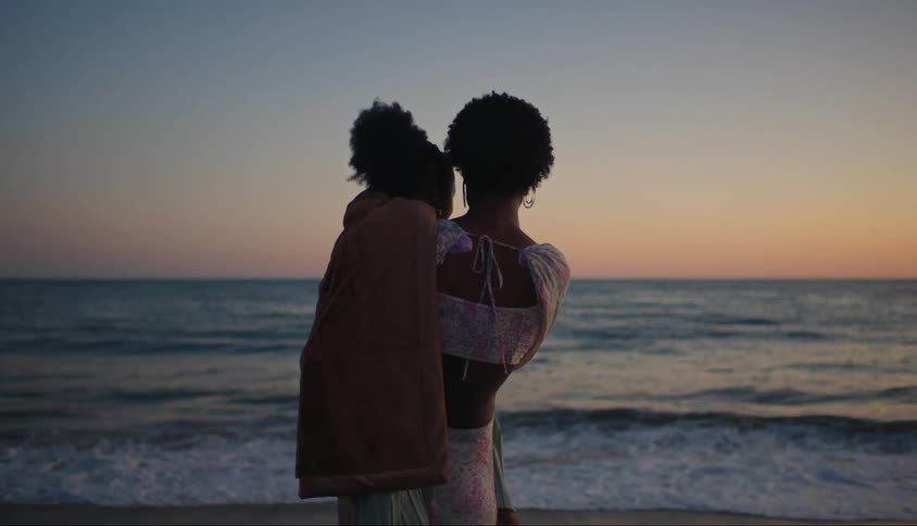 Mother with her daughter on her lap in front of the sea on the beach at sunset
