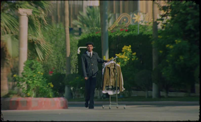 A bellboy pushing a clothing rack laden with various garments along a treelined sidewalk