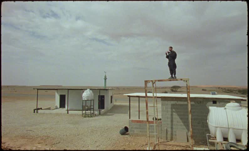 A man in black clothes stands atop a metal structure in the desert near a few buildings praying