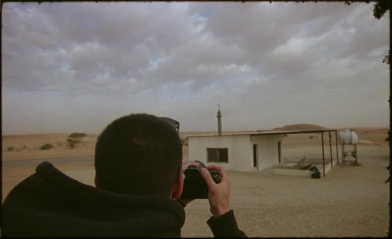 View of a man with a professional camera facing a small house in a desert environment
