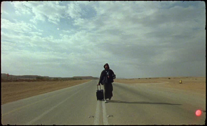 Man with sweatshirt and hood and a suitcase walking on a paved road in a desert environment