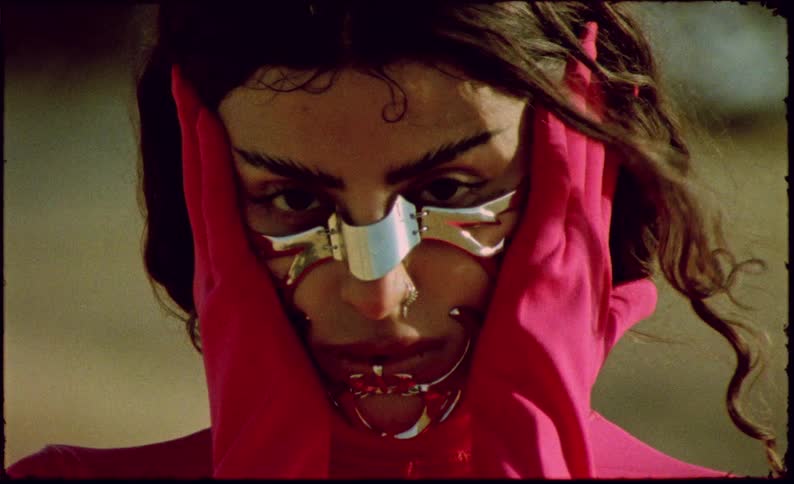 Close up of the face of a woman wearing a chromed metallic mask over her face wearing pink gloves and clothes
