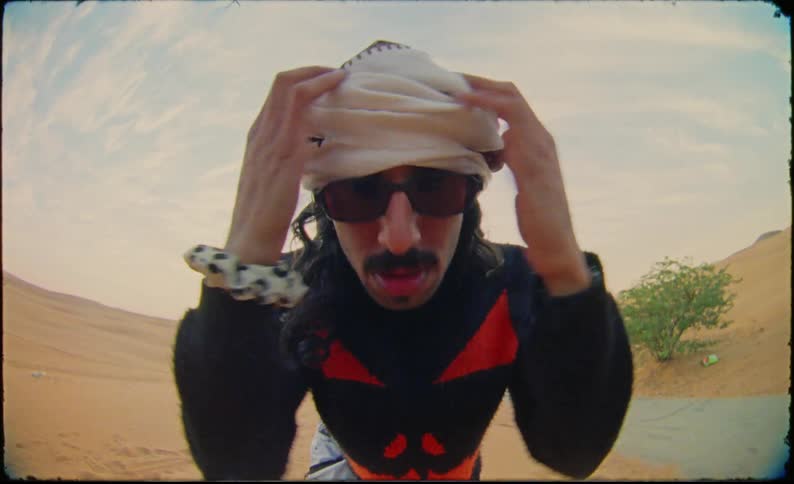 A man in a desert adjusts a white headscarf wearing a black garment with red accents in the desert