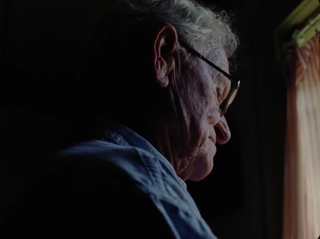 An old man wearing glasses stands by a window and looks outside