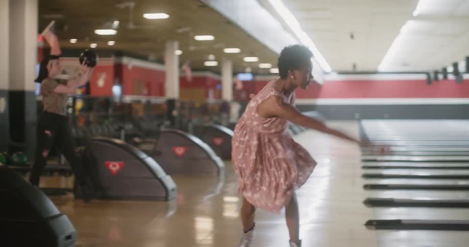 Two women in parallel bowling lanes throw their balls consecutively then one celebrates while the other is disappointed