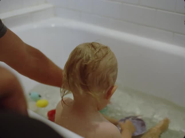 Father giving a bath to a baby who is playing in the bathtub with water and toys
