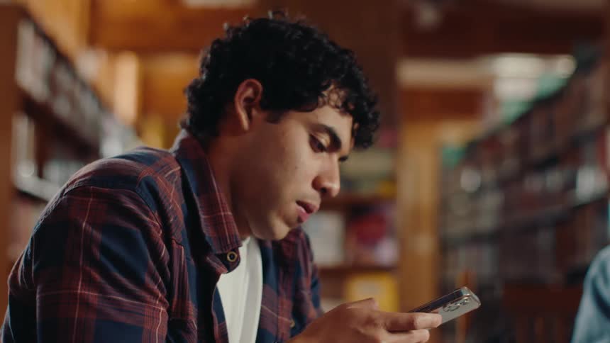 A man checks his cell phone and smiles in the library while friends study around him