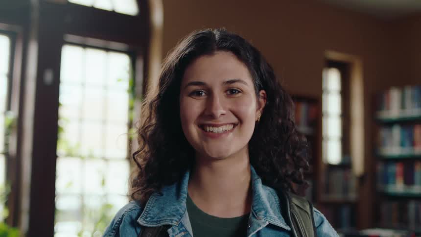 A portrait of a young female student with a backpack standing in the library while holding books smiling broadly