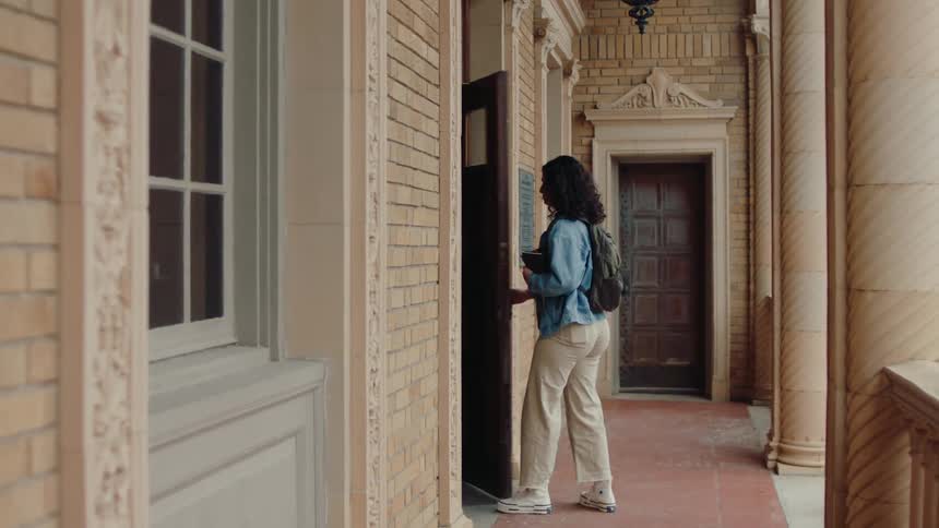 A young woman walks up to a library building and opens a door to enter it