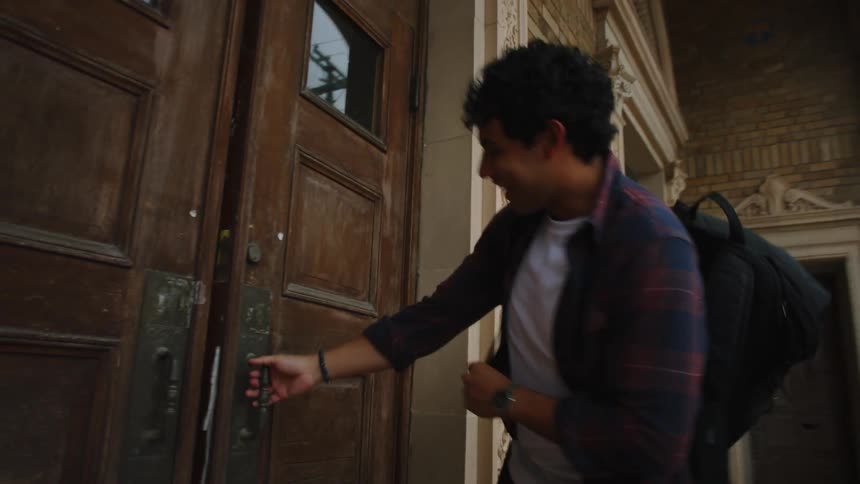 Two students wearing backpacks are chatting as they run up the stairs and enter an old columned library building with the male holding the door open with a smile as the female walks through.
