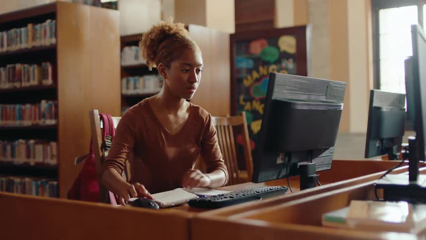 Woman takes notes while doing research on a computer at the library