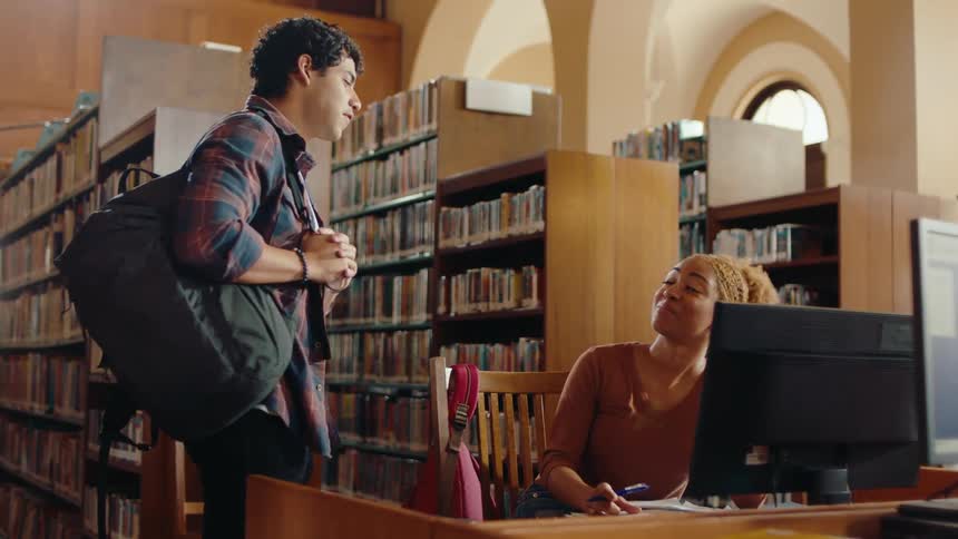 A young man walks into a library and meets a lady friend sitting at a desk with a computer and they talk and smile before he moves on