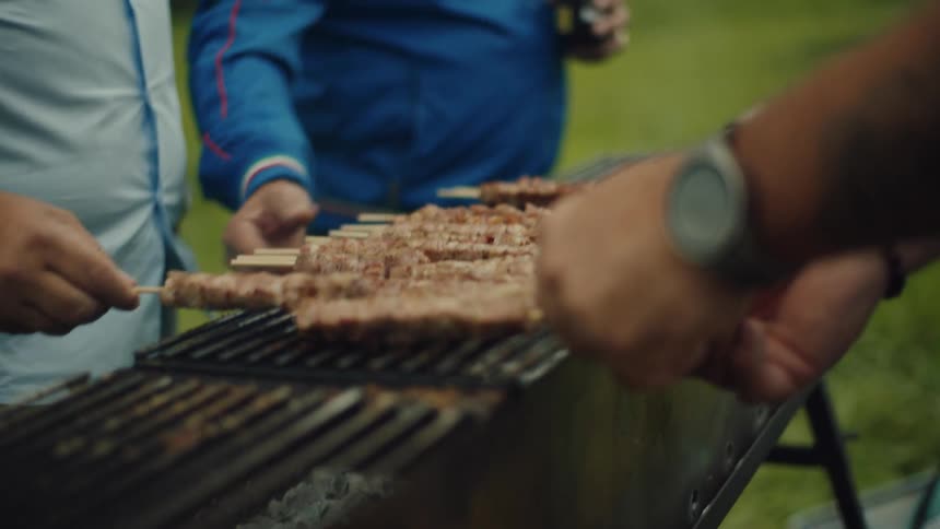 Hands of three men preparing meat skewers on a barbecue outdoors