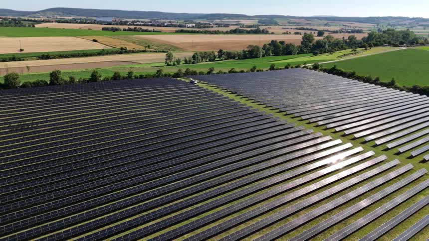 A field filled with solar panels sits in a rural area near farm fields