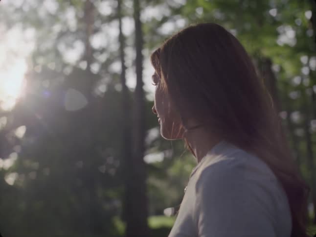 A young woman has her back looking at a forest full of trees and sunlight illuminating her face