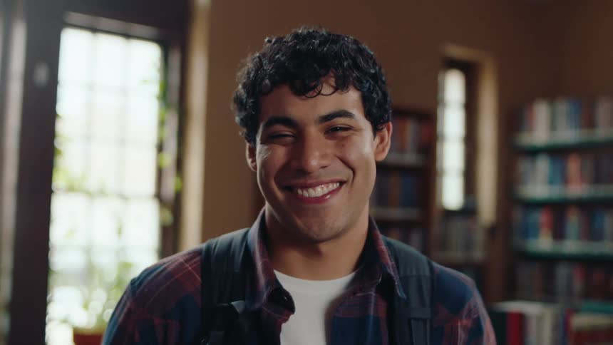 Portrait of a young latino male student with a backpack in a plaid shirt smiles while standing in a library near books