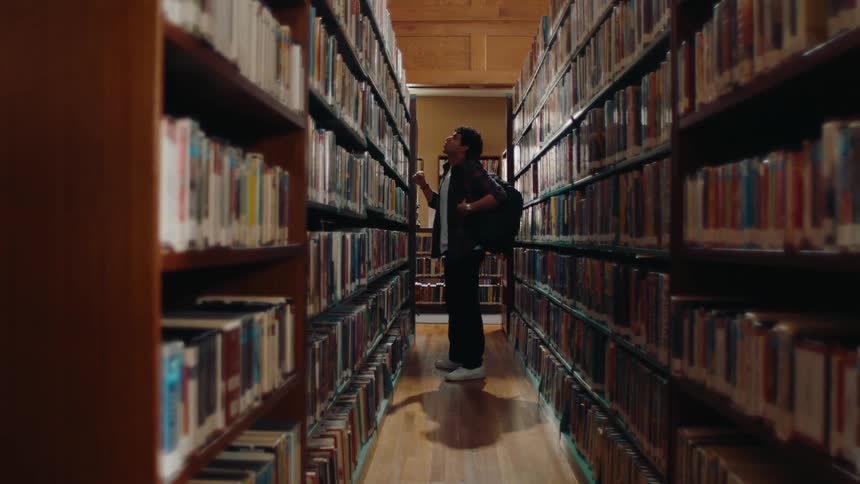 Latino student with backpack chooses a book from a bookshelf in a school library